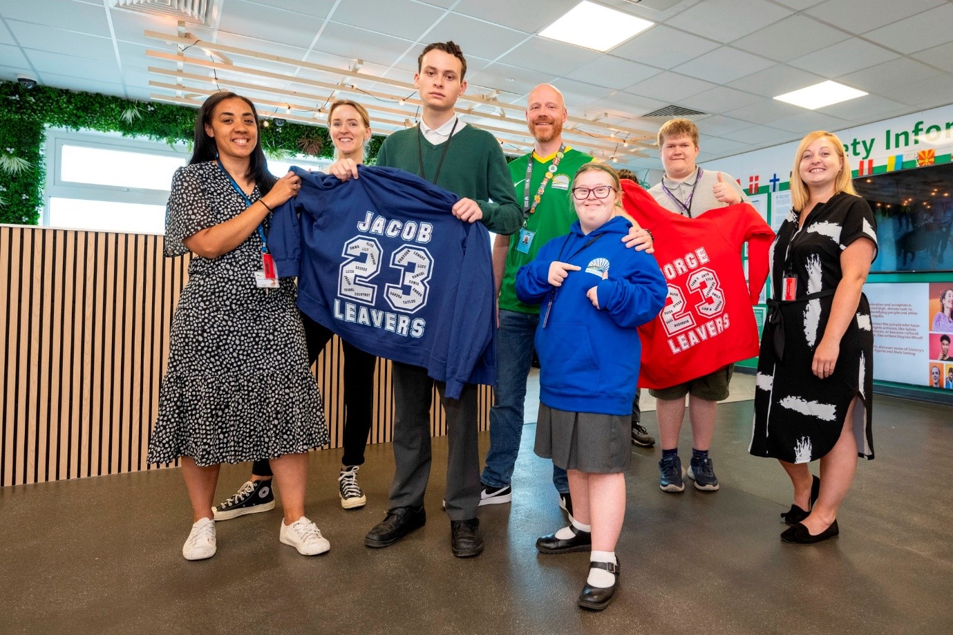 A group of seven people stands indoors. Two individuals in the center hold up sweatshirts. The left person holds a blue sweatshirt labeled “Jacob,” and the right person holds a red sweatshirt labeled “George.” Both sweatshirts feature the number “23” and the word “Leavers.” The background includes a wooden partition, some greenery.